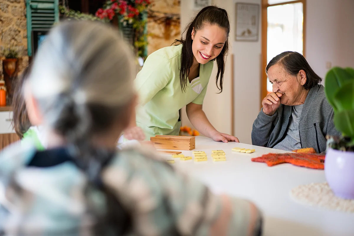 Caretaker assisting a group of laughing seniors playing a game of dominos